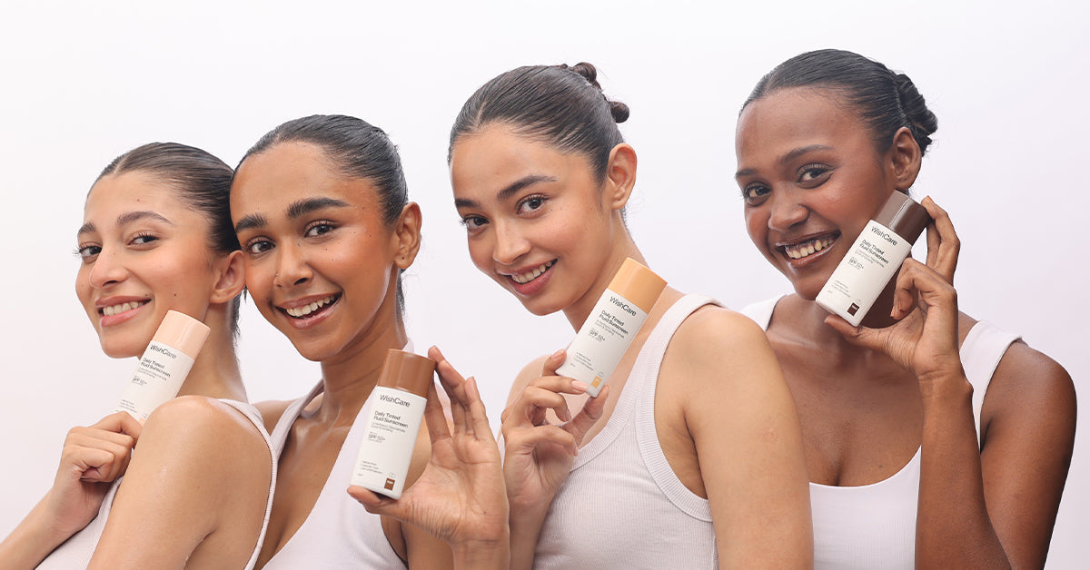 Four women with diverse Indian skin tones smiling and holding WishCare Daily Tinted Fluid Sunscreen bottles in shades ranging from light to deep, showcasing shade inclusivity for Indian skin tones.